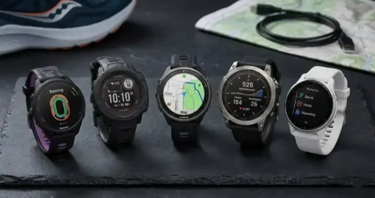 Diferentes modelos de relojes Garmin 2026 alineados, incluyendo Forerunner, Instinct, Fenix y Venu, sobre una mesa con material de entrenamiento y mapas.